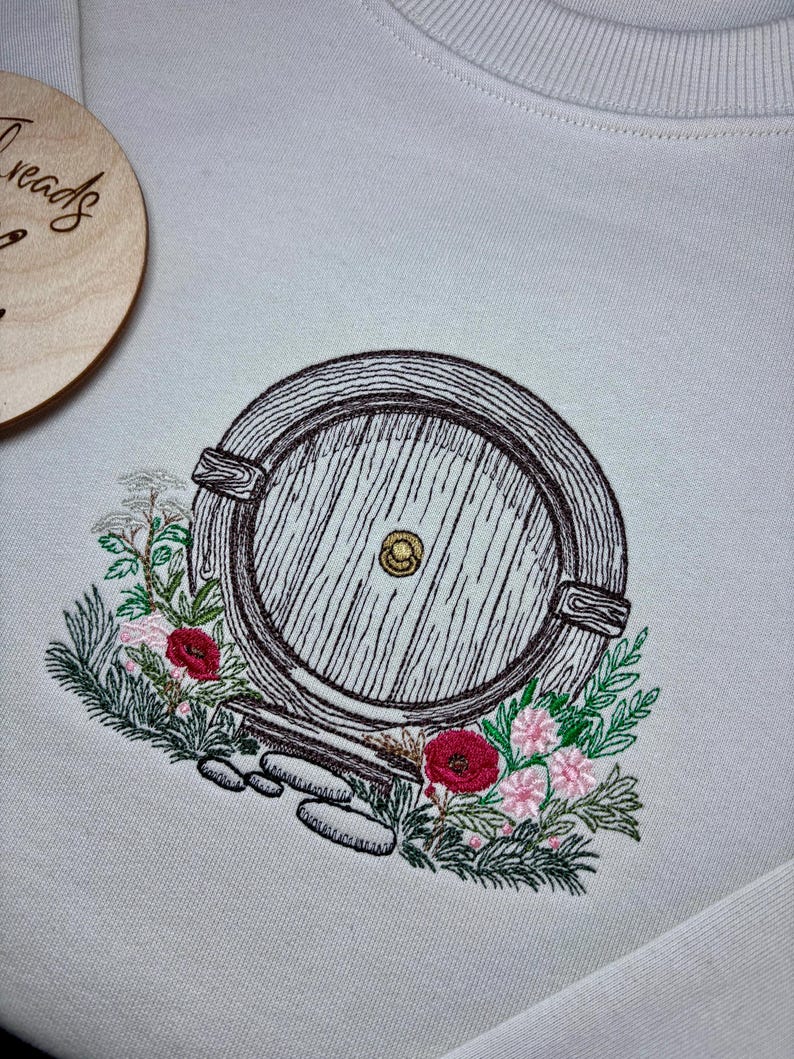 May include: A white sweatshirt with an embroidered image of a round wooden door with a knob, surrounded by green leaves and red and pink flowers.