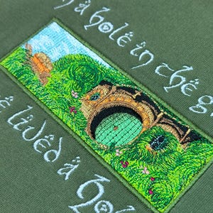 K&ouml;nnte beinhalten: Olivgr&uuml;nes Sweatshirt mit einem gestickten Aufn&auml;her, der eine Hobbit-Loch-Szene zeigt. Der Aufn&auml;her zeigt eine gr&uuml;ne T&uuml;r, &uuml;ppiges Gr&uuml;n und den Text "In a hole in the ground there lived a hobbit" in wei&szlig;er Schrift. Ein Holzschild ist zu sehen.