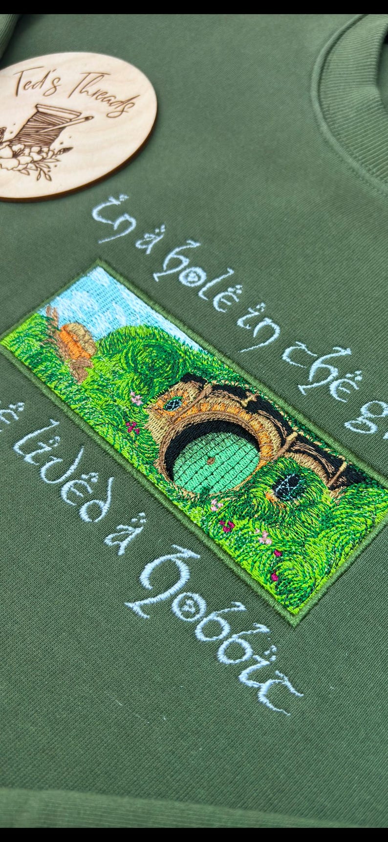 K&ouml;nnte beinhalten: Olivgr&uuml;nes Sweatshirt mit einem gestickten Aufn&auml;her, der eine Hobbit-Loch-Szene zeigt. Der Aufn&auml;her zeigt eine gr&uuml;ne T&uuml;r, &uuml;ppiges Gr&uuml;n und den Text "In a hole in the ground there lived a hobbit" in wei&szlig;er Schrift. Ein Holzschild ist zu sehen.