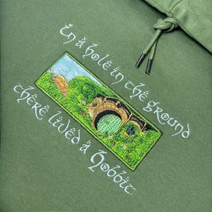 K&ouml;nnte beinhalten: Olivgr&uuml;ner Kapuzenpullover mit gesticktem Text und einem rechteckigen Aufn&auml;her mit einer Hobbit-Loch-Szene. Der Text lautet "In a hole in the ground there lived a hobbit" in wei&szlig;er Schrift. Der Hoodie hat einen Kordelzug.