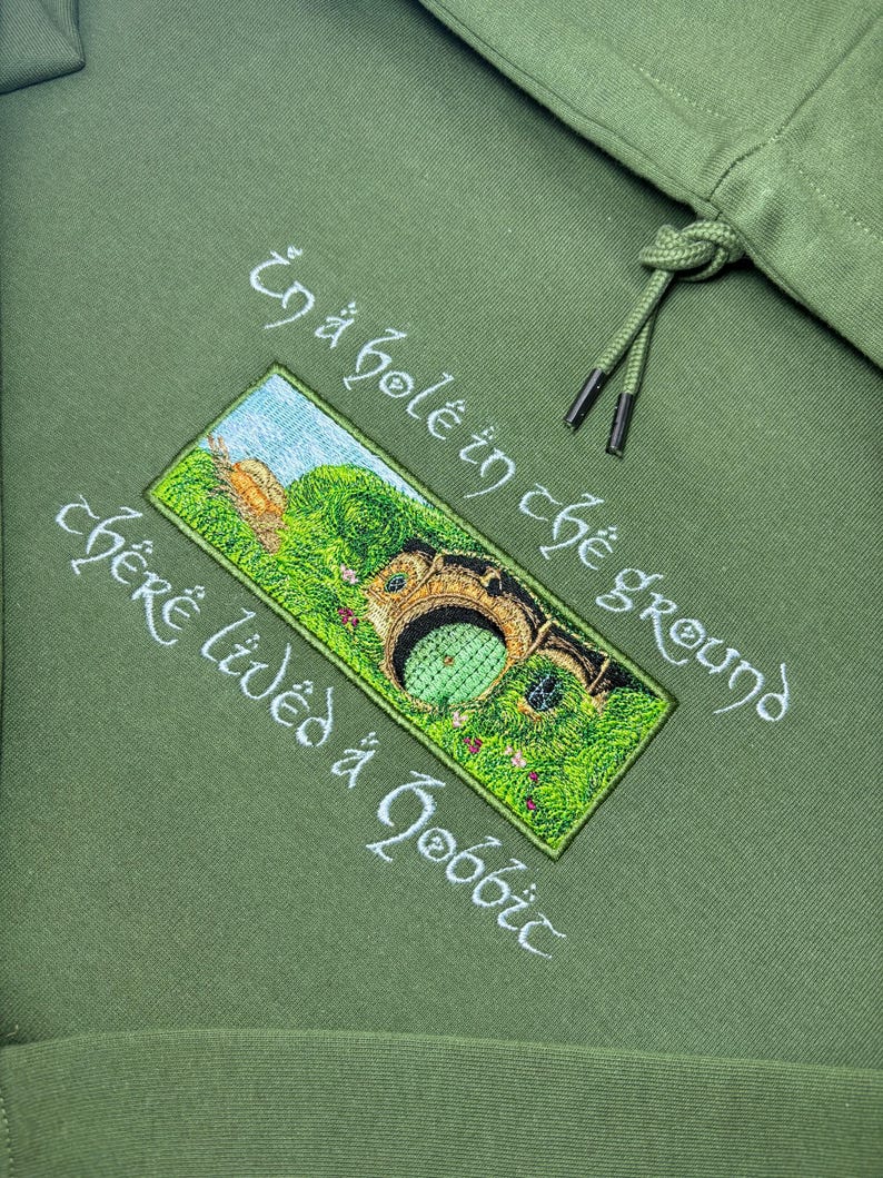 K&ouml;nnte beinhalten: Olivgr&uuml;ner Kapuzenpullover mit gesticktem Text und einem rechteckigen Aufn&auml;her mit einer Hobbit-Loch-Szene. Der Text lautet "In a hole in the ground there lived a hobbit" in wei&szlig;er Schrift. Der Hoodie hat einen Kordelzug.