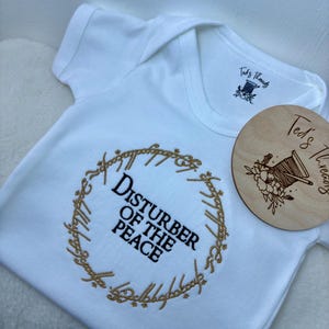 Puede incluir: Mono blanco para bebé con bordado dorado del Anillo Único de El Señor de los Anillos. La inscripción dice "Disturber of the Peace".