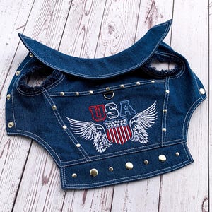 Può includere: Un giubbotto in denim blu per cani con un designo USA ricamato in bianco, rosso e blu con ali. Il giubbotto ha borchie argentate e un colletto.