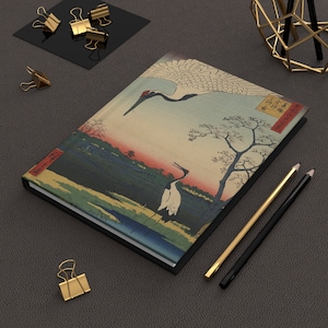 May include: A black and white notebook featuring a Japanese-style illustration of two cranes flying over a landscape. The notebook has a black spine and a textured cover.