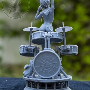 May include: A grey resin figurine of a drummer positioned behind a drum kit. The figure has short hair and wears headphones. The bass drum features a spiderweb design. The base is circular with a lattice structure. The text h3LLcreator is visible.