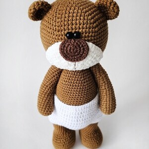 Pode incluir: Um urso de peluche de croch&ecirc; marrom vestindo uma fralda de croch&ecirc; branca. O urso tem olhos pretos e um nariz marrom.