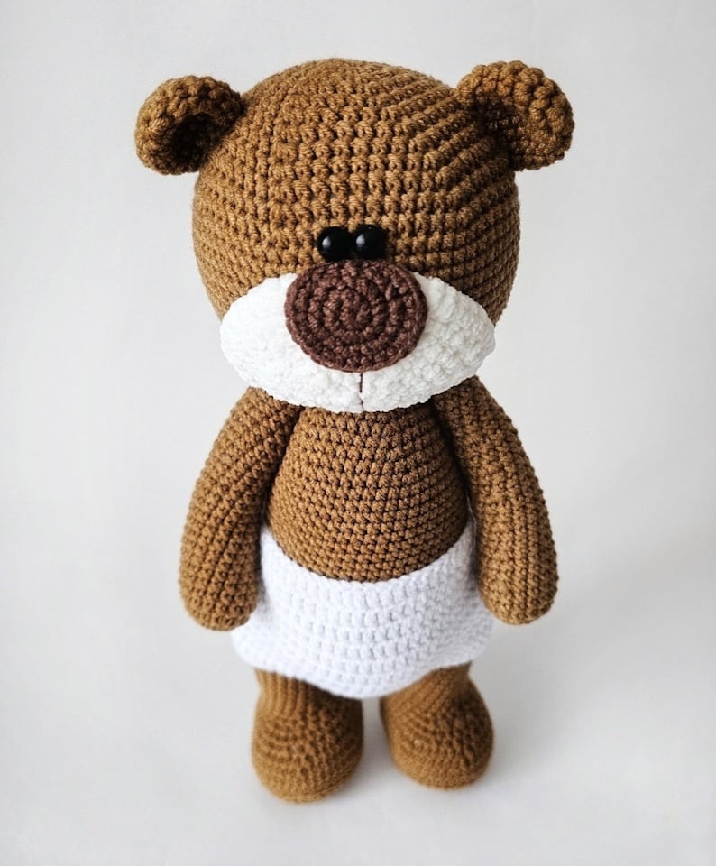 Pode incluir: Um urso de peluche de croch&ecirc; marrom vestindo uma fralda de croch&ecirc; branca. O urso tem olhos pretos e um nariz marrom.