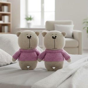 Puede incluir: Dos muñecos de osos de peluche tejidos. Los osos tienen cuerpos color beige, orejas pequeñas y visten suéteres rosas. Los osos tienen rasgos faciales cosidos en negro. Están sentados en una cama con ropa de cama blanca.