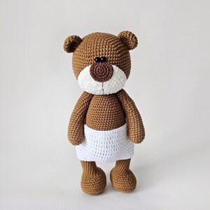 Pode incluir: Um urso de peluche de croch&ecirc; marrom vestindo uma fralda de croch&ecirc; branca. O urso tem olhos pretos e um nariz marrom.