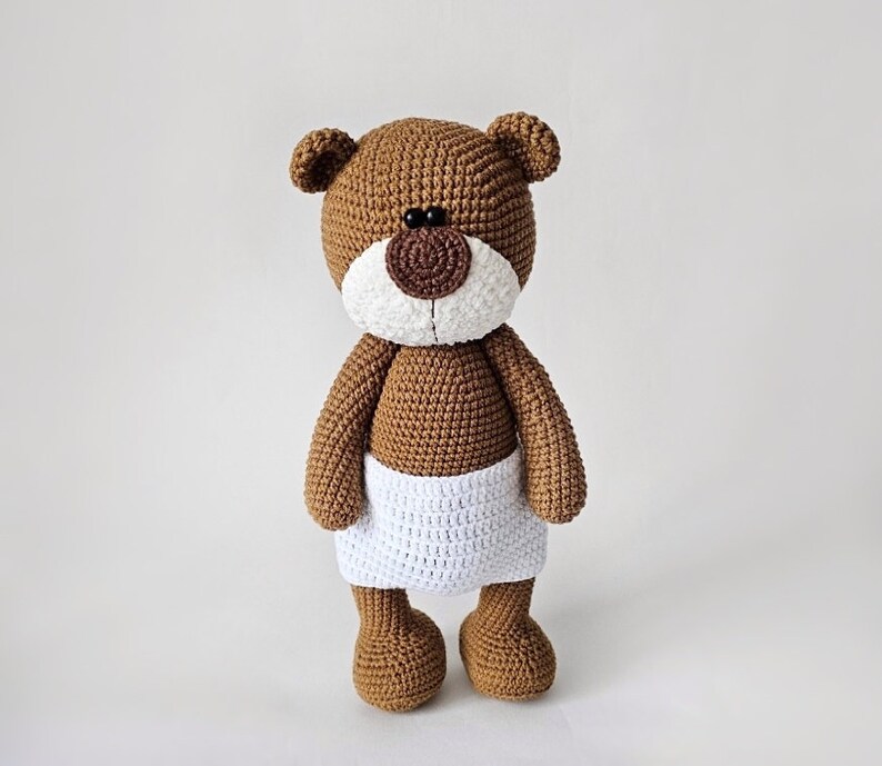 Pode incluir: Um urso de peluche de croch&ecirc; marrom vestindo uma fralda de croch&ecirc; branca. O urso tem olhos pretos e um nariz marrom.