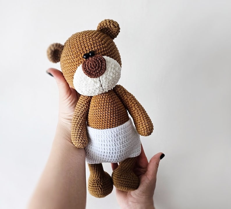 Pode incluir: Um urso de peluche de croch&ecirc; marrom vestindo uma fralda de croch&ecirc; branca. O urso tem olhos pretos e um nariz marrom.