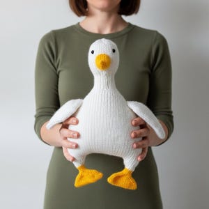 Könnte beinhalten: Ein weißes, gestricktes Plüschtier in Form einer Gans mit gelbem Schnabel und gelben Schwimmfüßen. Das Spielzeug hat einen runden Körper, zwei ausgestreckte Flügel und kleine schwarze Augen. Die Garnstruktur ist auf der Oberfläche sichtbar.