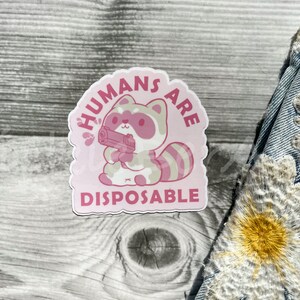 May include: Pink sticker with a cartoon raccoon holding a gun and the text "HUMANS ARE DISPOSABLE".