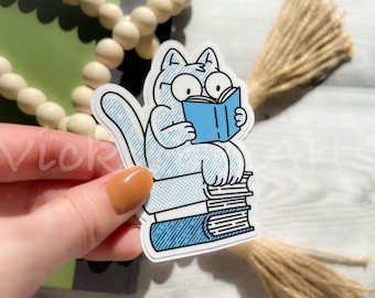 May include: A blue and white sticker of a cat reading a book while sitting on a stack of books. The cat has large eyes and a surprised expression.