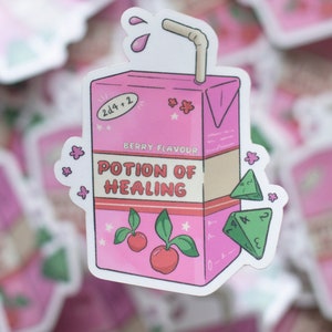 Puede incluir: Una pegatina de cartón de jugo de dibujos animados rosa y blanco con una pajita, dos cerezas y hojas verdes. La caja de jugo dice "Berry Flavour Potion of Healing" y tiene un dado verde en el lateral. La caja de jugo tiene un fondo blanco y acentos rosas.