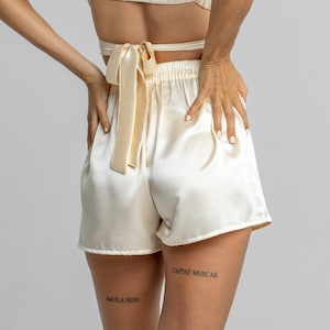 May include: A pair of cream-colored satin shorts with a matching satin top tied at the back. The shorts have a high waist and a relaxed fit. The model is wearing the shorts and top with a pair of tattoos on their legs that read "Captat Muscas" and "Aquila Non".