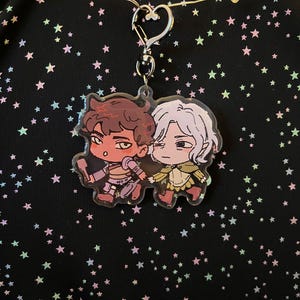 May include: A clear acrylic charm featuring two cartoon characters with brown and white hair, holding hands. The charm is attached to a silver-colored heart-shaped clasp. The background is black with colorful star shapes.