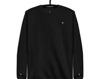 May include: Black crewneck sweatshirt with a small yellow embroidered logo on the chest and left sleeve. The sweatshirt is hanging on a black hanger. The logo is a stylized flame.