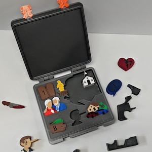 Puede incluir: Un estuche de rompecabezas gris con varias piezas de rompecabezas coloridas dispersas. Las piezas incluyen un corazón roto, una iglesia, una casa, un cuchillo y figuras. El estuche tiene dos clips naranjas en la parte superior.