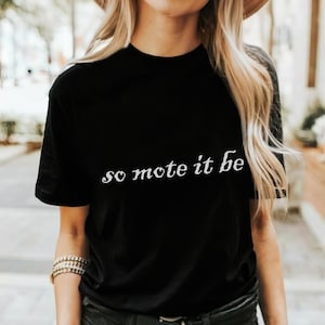 May include: Black t-shirt with the phrase "so mote it be" in white, stylized lettering. The shirt is a classic crew neck style.