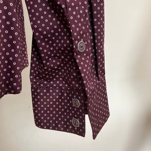 May include: Close-up of a maroon button-down shirt with a white polka dot pattern. The cuff features three silver buttons. The shirt's fabric is visible, showcasing the repeating dot design.