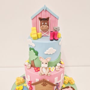 May include: A three-tiered farm-themed cake with a pink and blue color scheme. The top tier features a horse in a barn, the middle tier has a bunny, and the bottom tier includes a doghouse, a pig, and a pond with ducks.