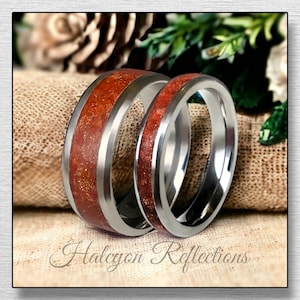 Puede incluir: Un par de anillos de boda con una banda de metal plateado y una incrustación roja y dorada. Los anillos están sobre una superficie de tela marrón.