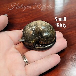 Pode incluir: Uma pequena figura de gato de cer&acirc;mica preta com glitter dourado embutido no esmalte. O gato est&aacute; enroscado em uma posi&ccedil;&atilde;o de dormir e tem o nome "Molly" inscrito no seu lado. A figura est&aacute; sendo segurada na m&atilde;o de uma pessoa.
