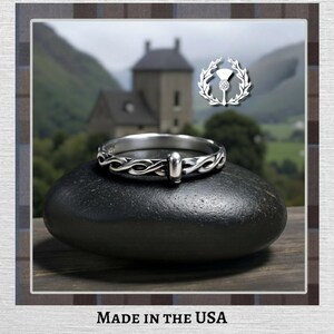 Puede incluir: Un anillo de plata con un intrincado diseño de nudo celta, apoyado sobre una piedra negra. El anillo presenta una barra central y una banda detallada. Un emblema de cardo es visible en el fondo, junto con una vista panorámica. El texto "MADE IN THE USA" está en la parte inferior.