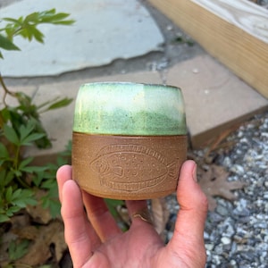 May include: A handmade ceramic tumbler with a brown base and a light green top. The brown section features an etched fish design. The tumbler is held in a hand, showcasing its size and craftsmanship. Ideal for beverages.