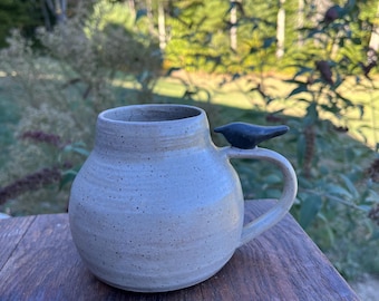 May include: A speckled, light gray ceramic mug with a rounded body and a dark blue bird-shaped handle. The mug is sitting on a wooden surface, with a blurred green and brown background.
