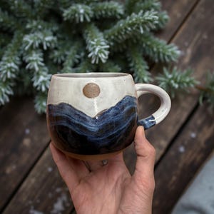 Puede incluir: Taza de cerámica hecha a mano con un diseño de montaña. La taza presenta una parte superior blanca con un sol marrón y un diseño de cordillera azul oscuro. El asa es blanca con un acento azul. La taza se sostiene en una mano.