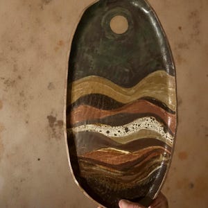 May include: An oval-shaped ceramic serving tray with a dark green top section featuring a circular cutout. The tray's design includes layered, wavy bands of brown, tan, and white, creating a textured, earthy aesthetic. The tray is held by a hand.