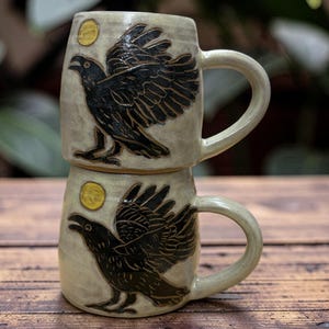 May include: Two stacked ceramic mugs with a raven design. The mugs are beige with a black raven illustration and a yellow moon. The mugs have a curved handle and are sitting on a wooden surface.