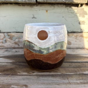 May include: A handmade ceramic mug with a landscape design. The mug features a white top with a brown sun, green and brown bands, and a dark brown base. The mug is sitting on a wooden surface.