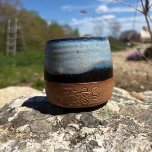 May include: A handmade ceramic tumbler with a blue and black glazed top and a brown base. The base features an etched fish design. The tumbler is sitting on a rock outdoors with a blurred background of trees and sky.