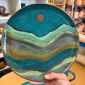 May include: A handmade ceramic plate with a teal and brown color scheme. The plate features a landscape design with layered green and blue tones, and a brown circle in the upper center. The plate has a brown rim.