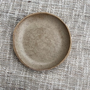 May include: A handmade, speckled ceramic plate with a slightly raised rim. The plate is a neutral beige color with darker speckles throughout. The plate is round and appears to be approximately 8 inches in diameter.