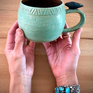 Pode incluir: Caneca de cerâmica artesanal com esmalte verde claro. A caneca apresenta uma faixa decorativa de triângulos esculpidos perto da borda e um detalhe azul escuro na alça. A caneca é segurada por duas mãos, mostrando uma pulseira turquesa.