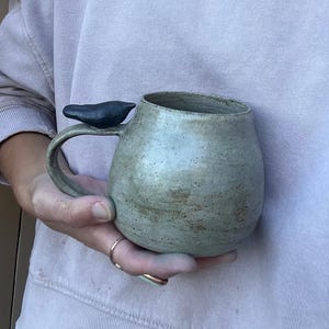 May include: A handcrafted ceramic mug with a distinctive bird-shaped handle. The mug features a speckled, earthy green glaze and a dark grey bird detail. The mug is held in a person's hands, highlighting its size and design. A ring is visible on the person's finger.