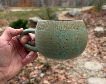 Puede incluir: Una taza de cerámica hecha a mano con un cuerpo redondeado y un asa. La taza es de color verde azulado moteado con un borde texturizado marrón claro. La taza se sostiene en una mano, con un fondo borroso al aire libre.