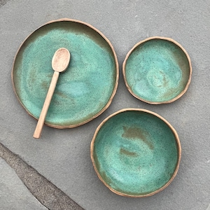 May include: A handmade ceramic tableware set featuring a large plate, two bowls, and a wooden spoon. The set has a turquoise glaze with brown rims. The pieces are arranged on a gray surface.