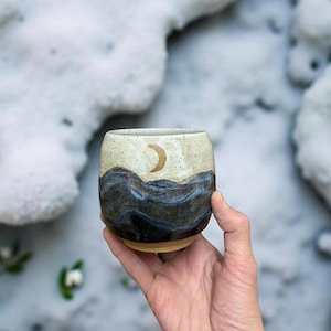 Peut inclure: Un gobelet en céramique fait à la main avec un motif de paysage. La partie supérieure est beige moucheté, passant à un motif de vagues bleu foncé et marron. Un croissant de lune doré est peint sur la section beige. Le gobelet est tenu devant un fond enneigé.