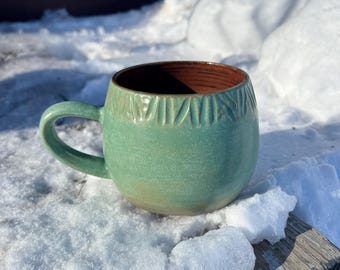 Puede incluir: Una taza de cerámica hecha a mano con forma redondeada y un esmalte verde. La taza tiene un interior marrón y un asa. Líneas decorativas talladas rodean la parte superior. La taza está sobre una superficie de madera cubierta de nieve.