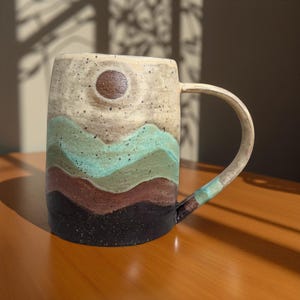 May include: A handmade ceramic mug with a landscape design. The mug features a speckled beige background with a brown sun and layered mountain design in shades of green, brown, and black. The handle and rim are beige. The mug sits on a wooden surface.