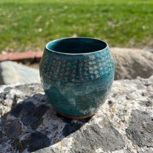 May include: A handcrafted teal ceramic tumbler with a textured surface. The small, round vessel has a wide opening and a speckled pattern around the top. The base is a natural, unglazed clay color. The tumbler sits on a gray rock in natural light.