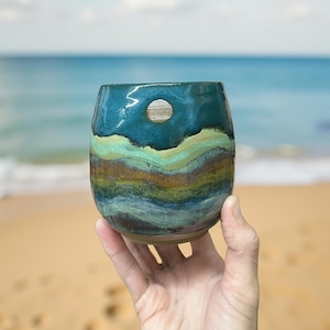 Op de afbeelding: Een handgemaakte keramische tumbler met een uniek, gelaagd ontwerp. De tumbler heeft een donkerblauwe bovenkant, die overgaat in banden van groen, geel en bruin. Een klein, cirkelvormig houten accent is in de rand gezet. De tumbler wordt tegen een achtergrond van een strand en oceaan gehouden.