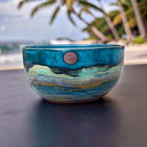 May include: A handcrafted ceramic bowl with a turquoise glaze and a brown circular accent. The bowl features layered bands of blue, green, and gold, evoking a coastal landscape. The bowl is approximately 5 inches in diameter.