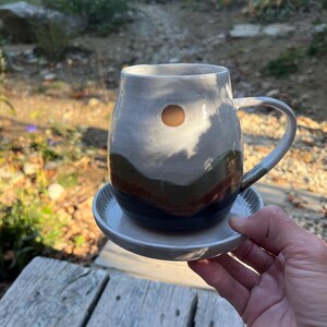May include: Handmade ceramic mug with a matching saucer. The mug features a unique design with a brown circle and layered earth-tone colors. The handle is curved, and the saucer has a ridged edge. The mug is held over a wooden surface.