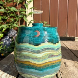 May include: A ceramic jar with a lid, featuring a glossy teal upper section and a multi-colored wavy pattern in shades of light green, yellow, and blue across the body. A small, reddish-brown crescent moon symbol is centered near the top rim.